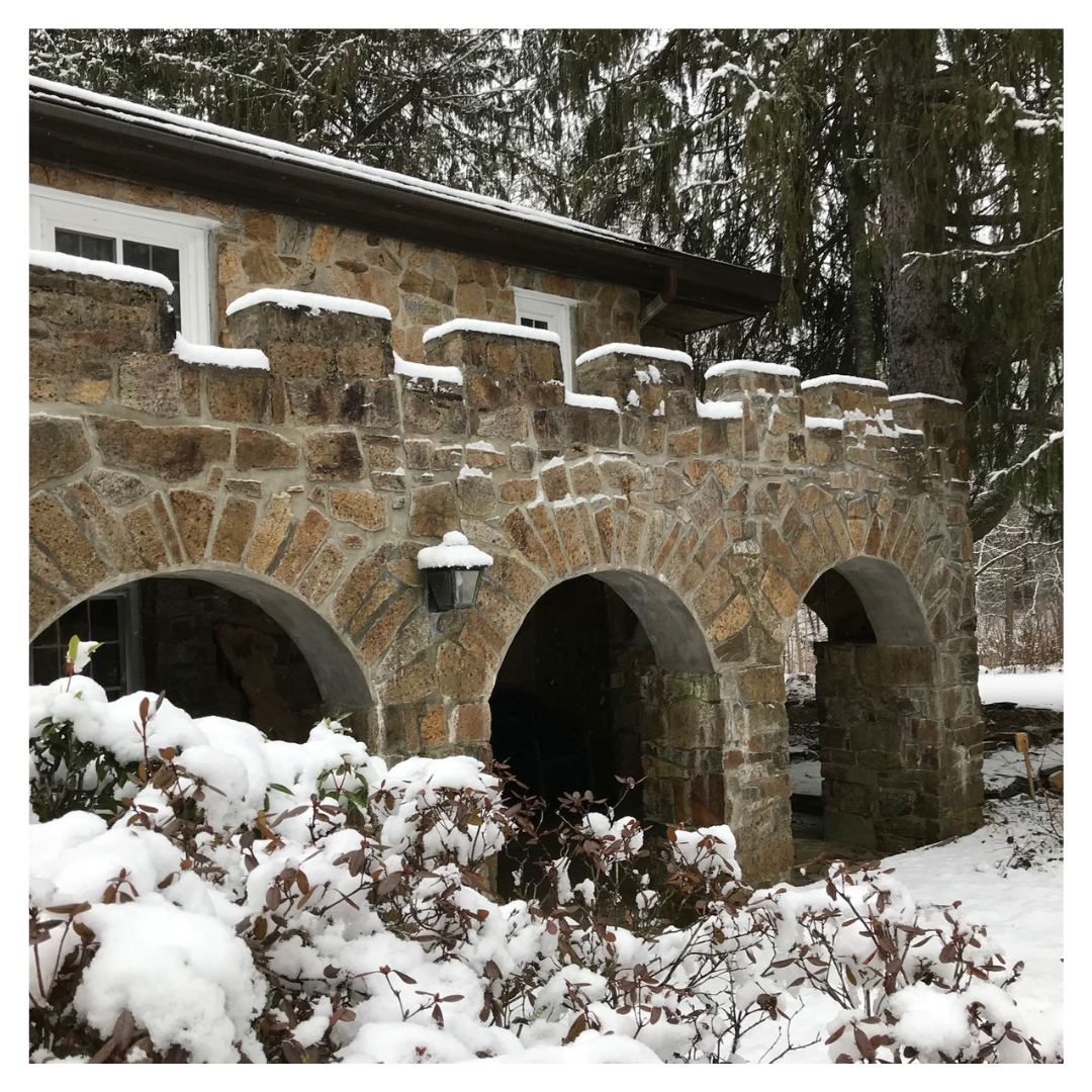 Snow covers evergreen trees and a building with stone arches framing a portico